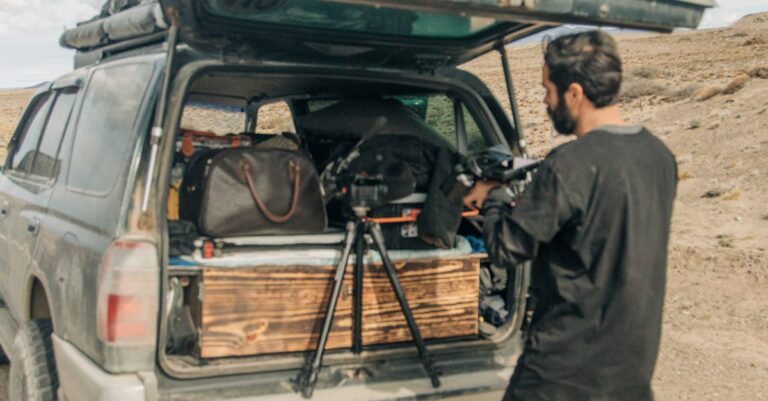 A man prepares his camera gear from the back of an SUV in a desert landscape.