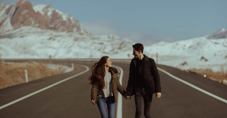 A romantic couple walking hand in hand on a scenic road with snowy mountains in the background.