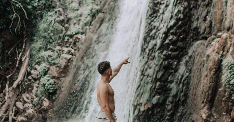 Shirtless man standing by a waterfall, appreciating nature's beauty.