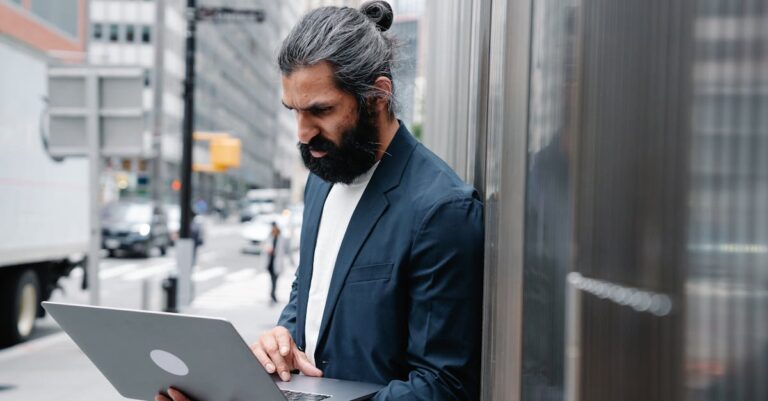 Bearded businessman focusing on laptop work in an urban outdoor setting.