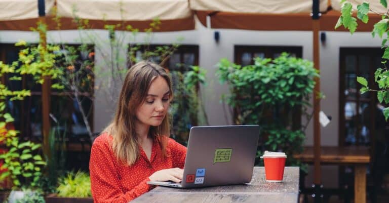 Young woman working on a laptop in an outdoor cafe setting, enjoying a cup of coffee.